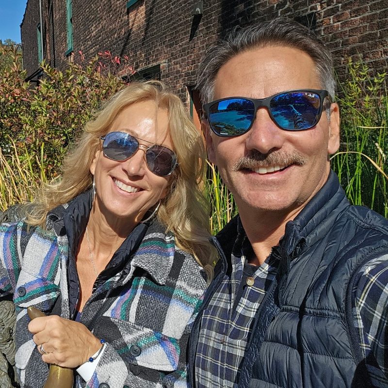 Joe, on the right, and Dar smiling in sunglasses outside a brick building on a sunny day